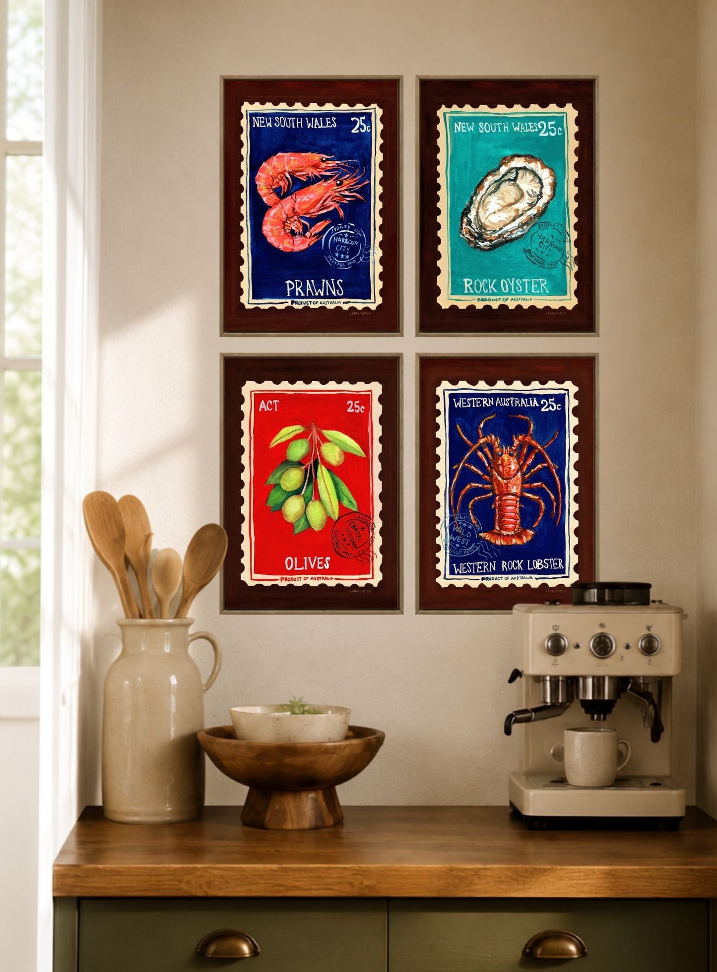 Four framed vintage-style seafood postage stamp prints arranged in a 2x2 grid on a beige wall above a wooden countertop with a ceramic pitcher, bowl, and espresso machine.