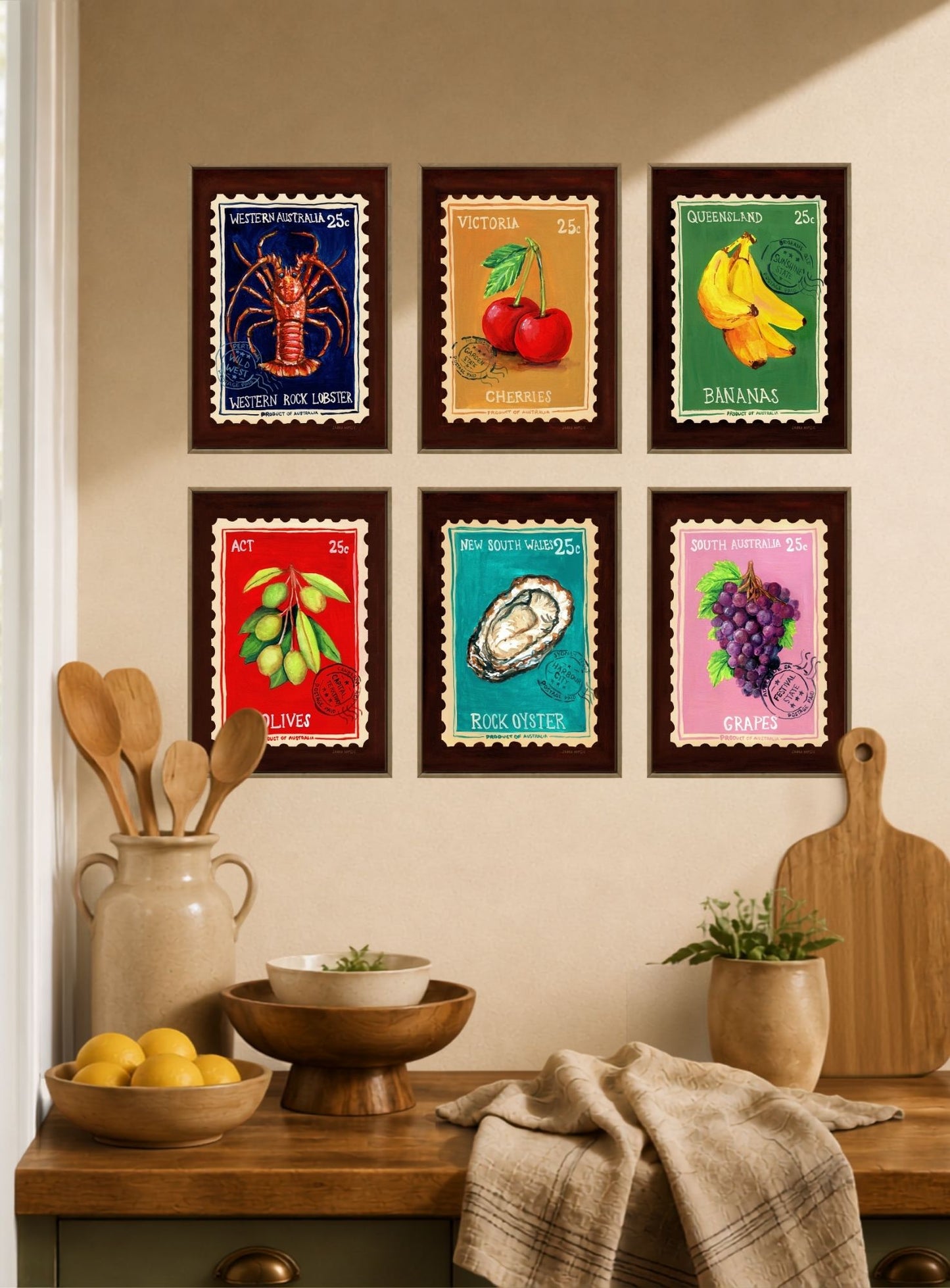 Six framed colorful art prints depicting seafood and fruit illustrations are arranged in a 2x3 grid on a beige wall above a wooden kitchen counter with bowls, lemons, and a cutting board.