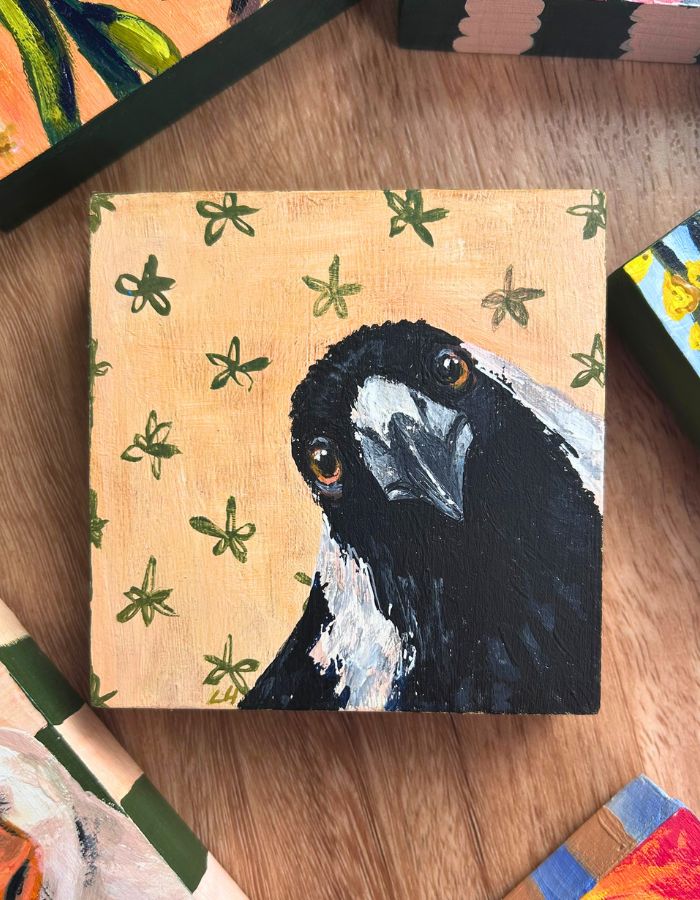 Original Australian magpie painting by Laura Hamzic, featuring a close-up magpie portrait on a cream background with green floral motifs, photographed on a timber surface.