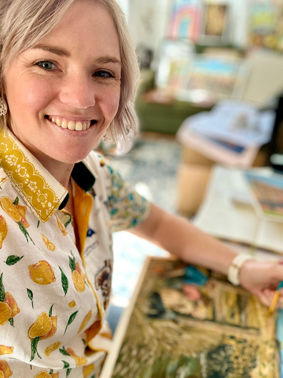 A smiling woman with short blonde hair wearing a white short-sleeve button-up shirt with a yellow lemon print, holding a colorful painting in a brightly lit room.