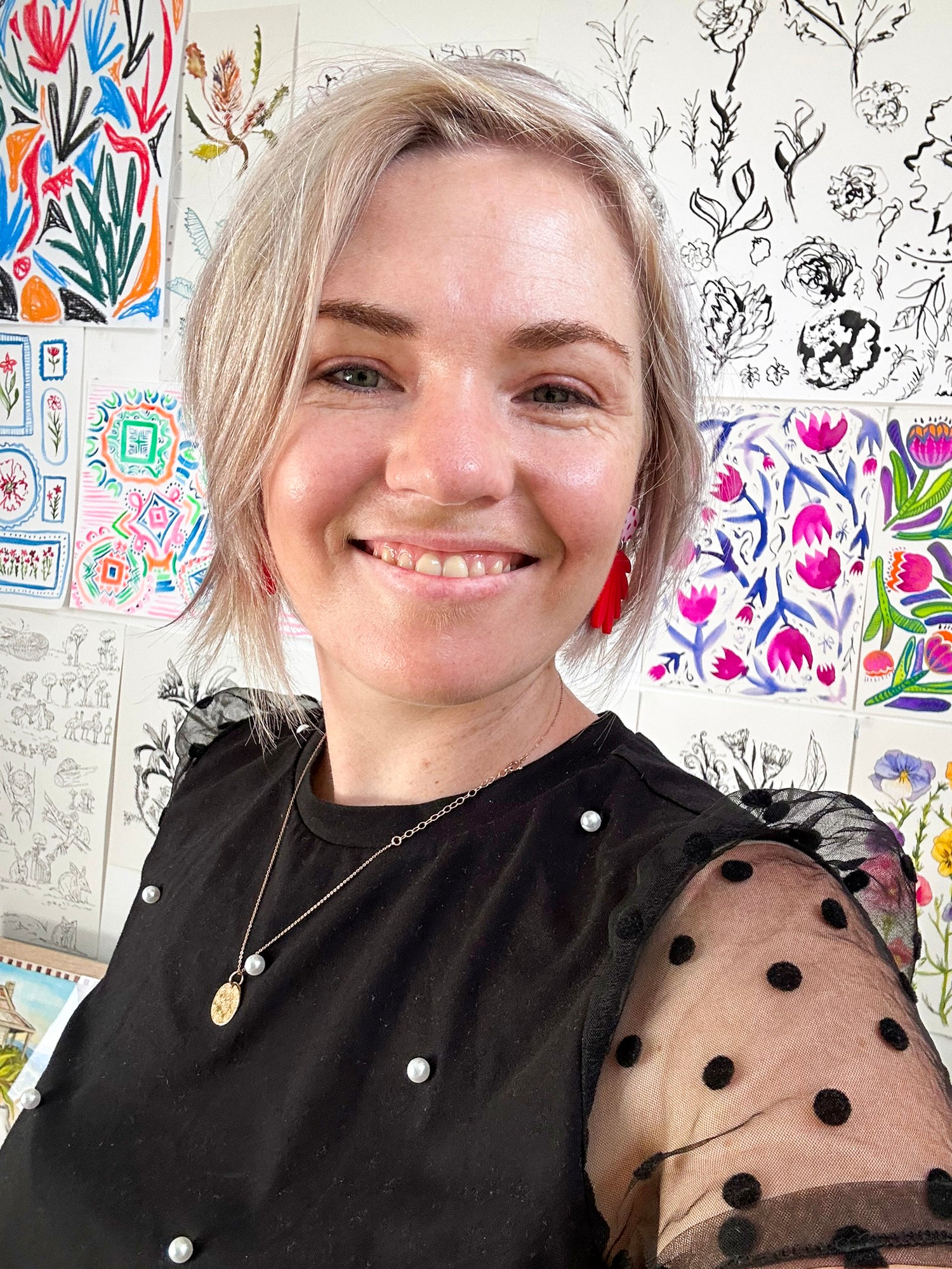 Laura Hamzic - Brisbane artist and surface pattern designer with blonde hair and red earrings smiling in front of a colorful wall with artistic designs.