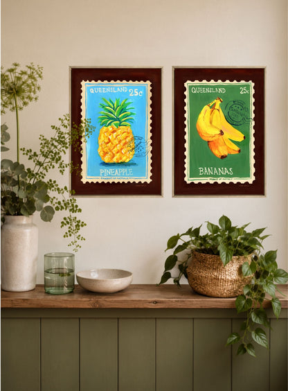 Two framed art prints featuring vintage Queensland postage stamps with a pineapple and bananas, displayed on a wall above a wooden shelf with plants and home decor.