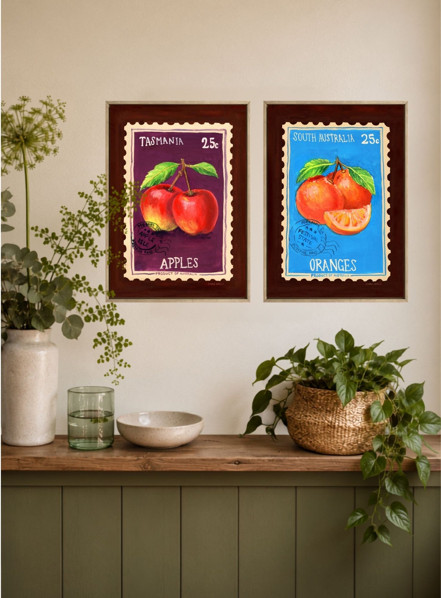 Two framed vintage-style Australian fruit posters hang on a beige wall above a wooden shelf with plants and decorative items. The left poster shows red apples on a purple background labeled 'TASMANIA 2½', and the right poster shows oranges on a blue background labeled 'SAUTERELAND 2½'.