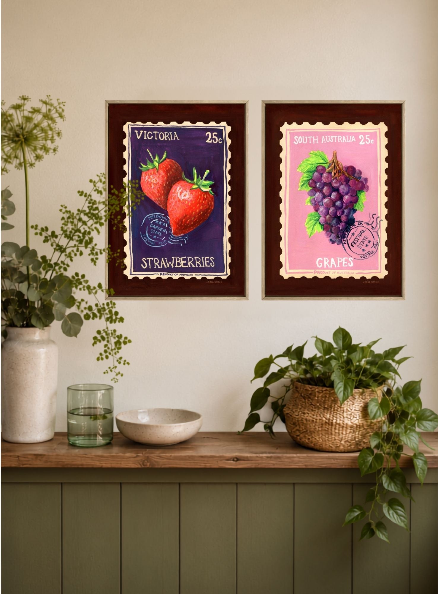 Two framed vintage-style fruit posters hang on a beige wall above a wooden console table with plants and decor. The left poster shows strawberries in a purple stamp design labeled Victoria 25c, and the right poster shows grapes in a pink stamp design labeled South African 25c.