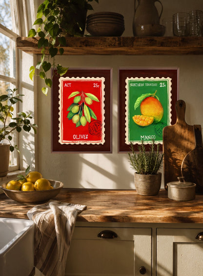 Two vintage-style framed fruit posters hanging on a kitchen wall above a wooden counter. The left poster shows green olives on a red background labeled 'OLIVES', and the right poster shows a mango on a green background labeled 'MANGO'.