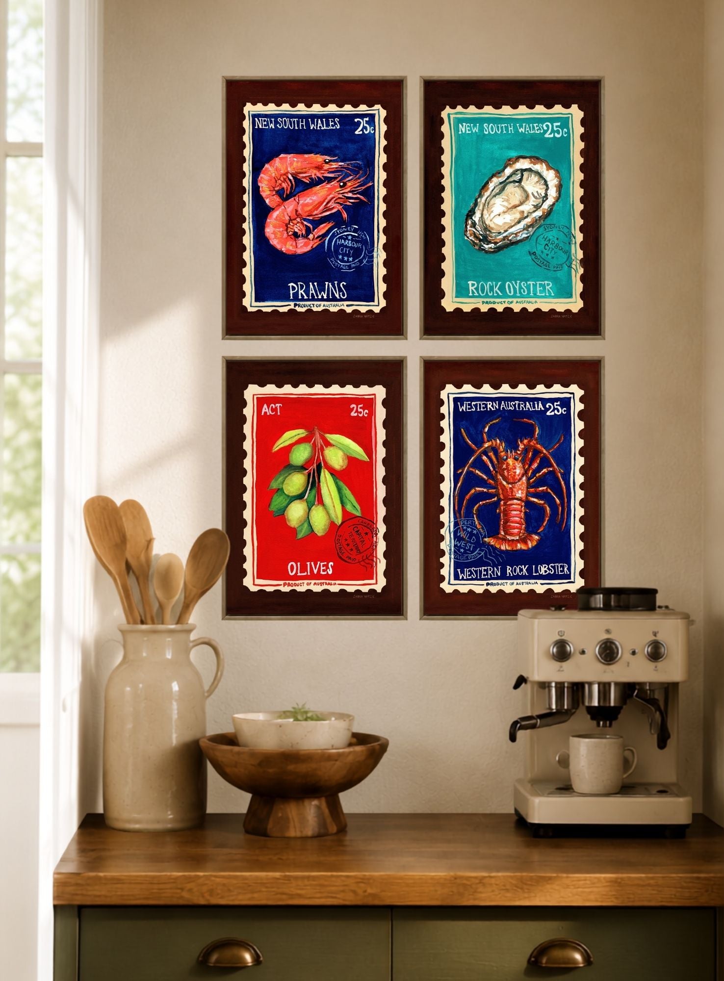 Four framed vintage-style seafood postage stamp prints arranged in a 2x2 grid on a beige wall above a wooden countertop with a ceramic pitcher, bowl, and espresso machine.