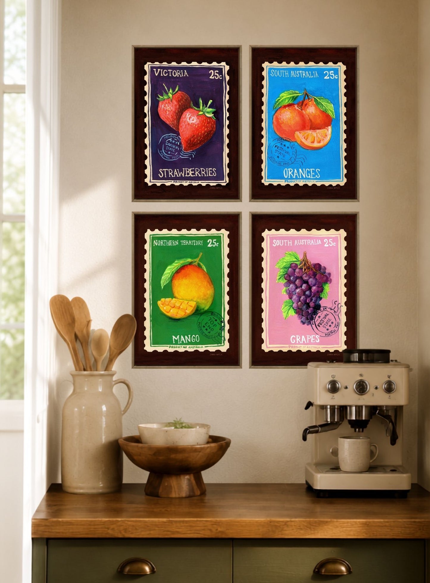 Four framed fruit art prints are displayed on a beige wall above a wooden kitchen counter. The prints feature colorful illustrations of strawberries, oranges, mango, and grapes in vintage stamp-style designs.