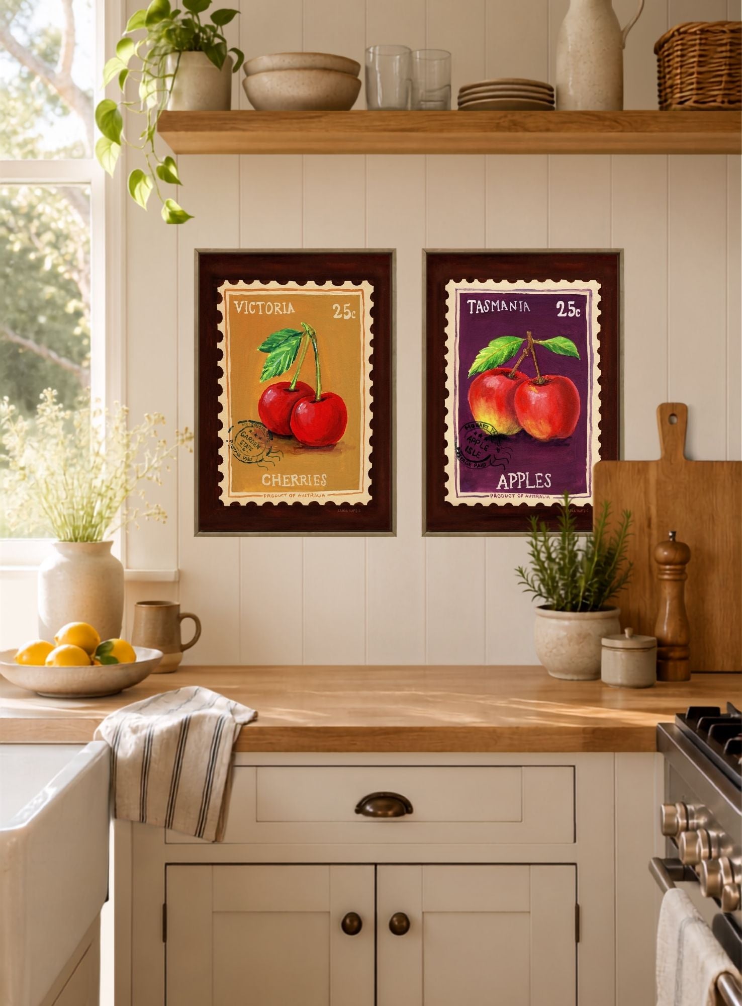 Two framed vintage-style fruit art prints hanging on a white paneled kitchen wall above a wooden countertop. The left print shows red cherries with the text 'VICTORIA 25C CHERRIES', and the right print shows red apples with the text 'TASMANIA 25C APPLES'.