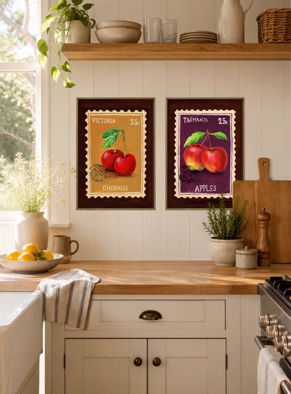 Two framed vintage-style fruit art prints hanging on a white paneled kitchen wall above a wooden countertop. The left print shows red cherries with the text 'VICTORIA 25C CHERRIES', and the right print shows red apples with the text 'TASMANIA 25C APPLES'.