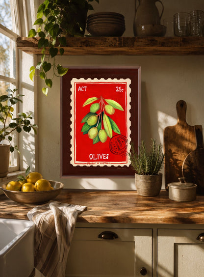A framed vintage-style art print of olives on a red background hangs on a kitchen wall above a wooden countertop with plants, lemons, and kitchen items.