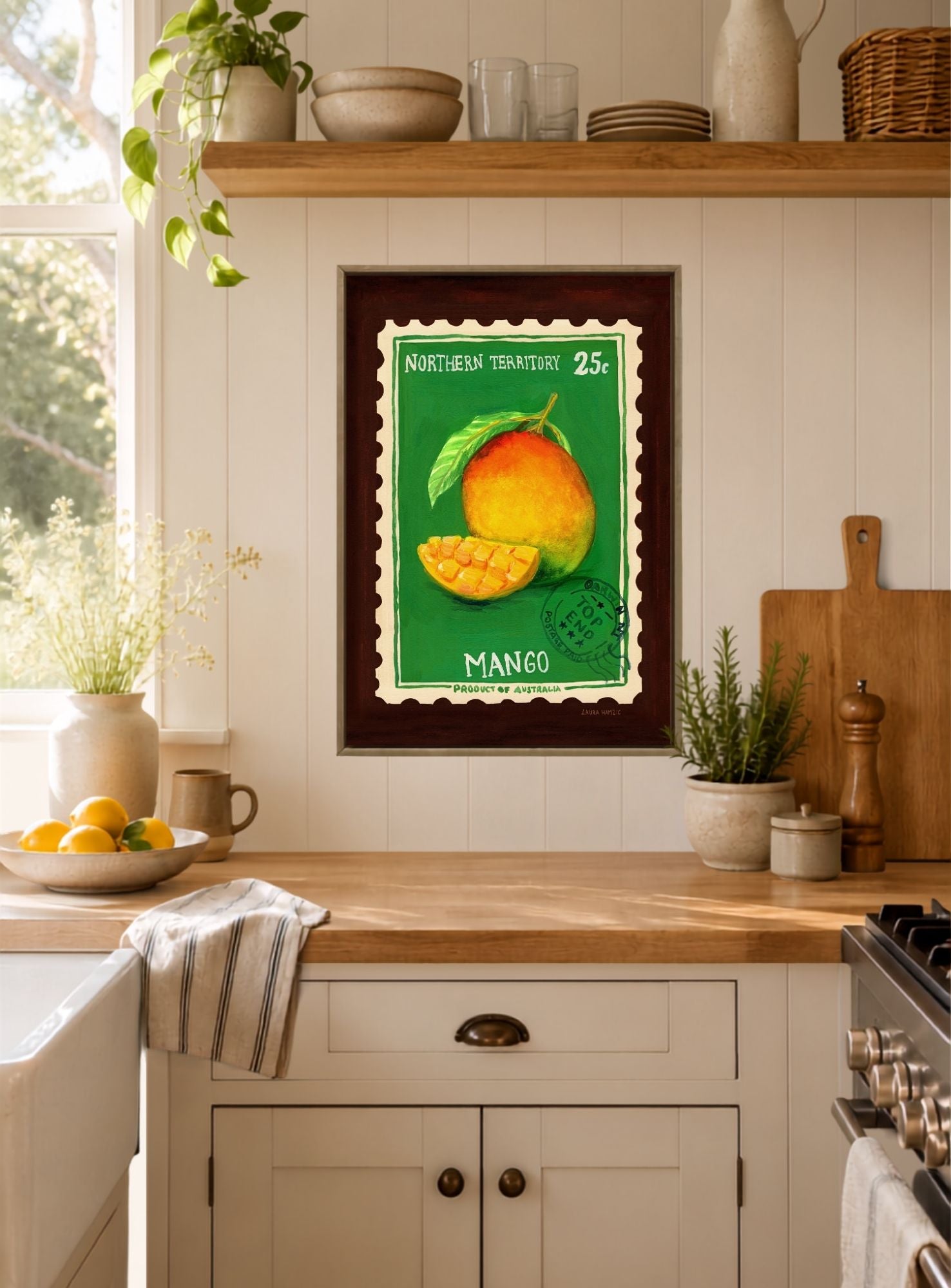 A framed vintage-style mango poster hangs on a white paneled kitchen wall above a wooden countertop. The print shows a green background with a yellow-orange mango and the word 'MANGO,' while the surrounding kitchen includes plants, bowls, and a stove.
