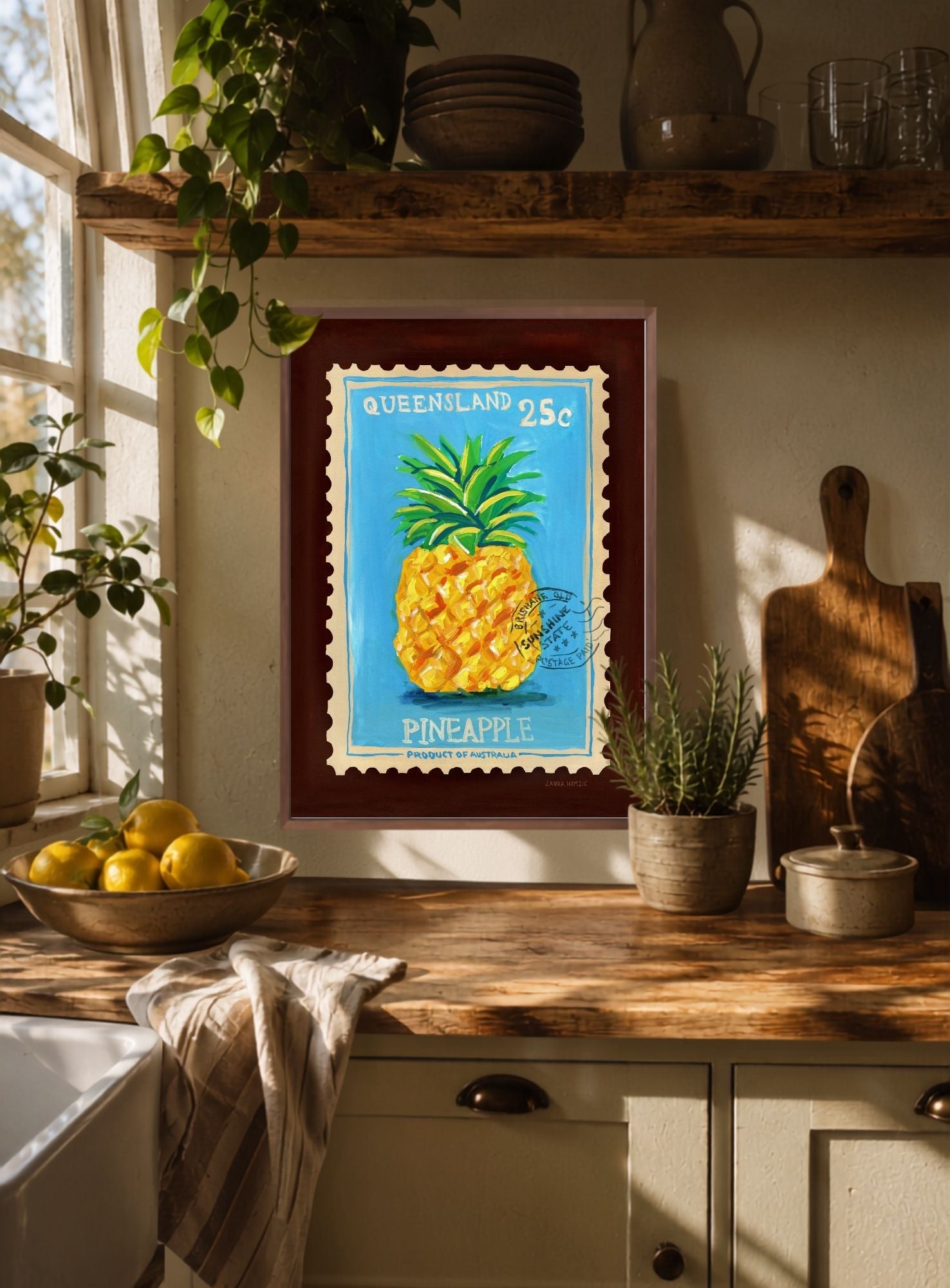 A framed art print featuring a retro-style pineapple stamp design with the text 'QUEENSLAND 25c PINEAPPLE' is displayed on a kitchen wall above a wooden countertop with plants and lemons.