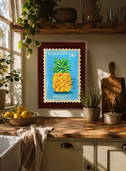 A framed art print featuring a retro-style pineapple stamp design with the text 'QUEENSLAND 25c PINEAPPLE' is displayed on a kitchen wall above a wooden countertop with plants and lemons.