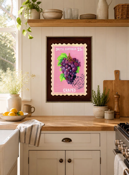 A framed art print featuring a vintage South Australian postage stamp design with purple grapes on a pink background, displayed on a kitchen wall above a wooden countertop.