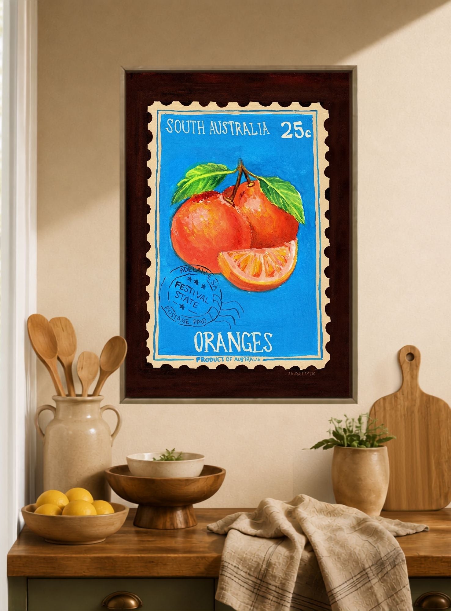 A framed art print of a vintage South Australia postage stamp featuring oranges, styled on a beige wall above a wooden kitchen counter with lemons, wooden utensils, and a cutting board.