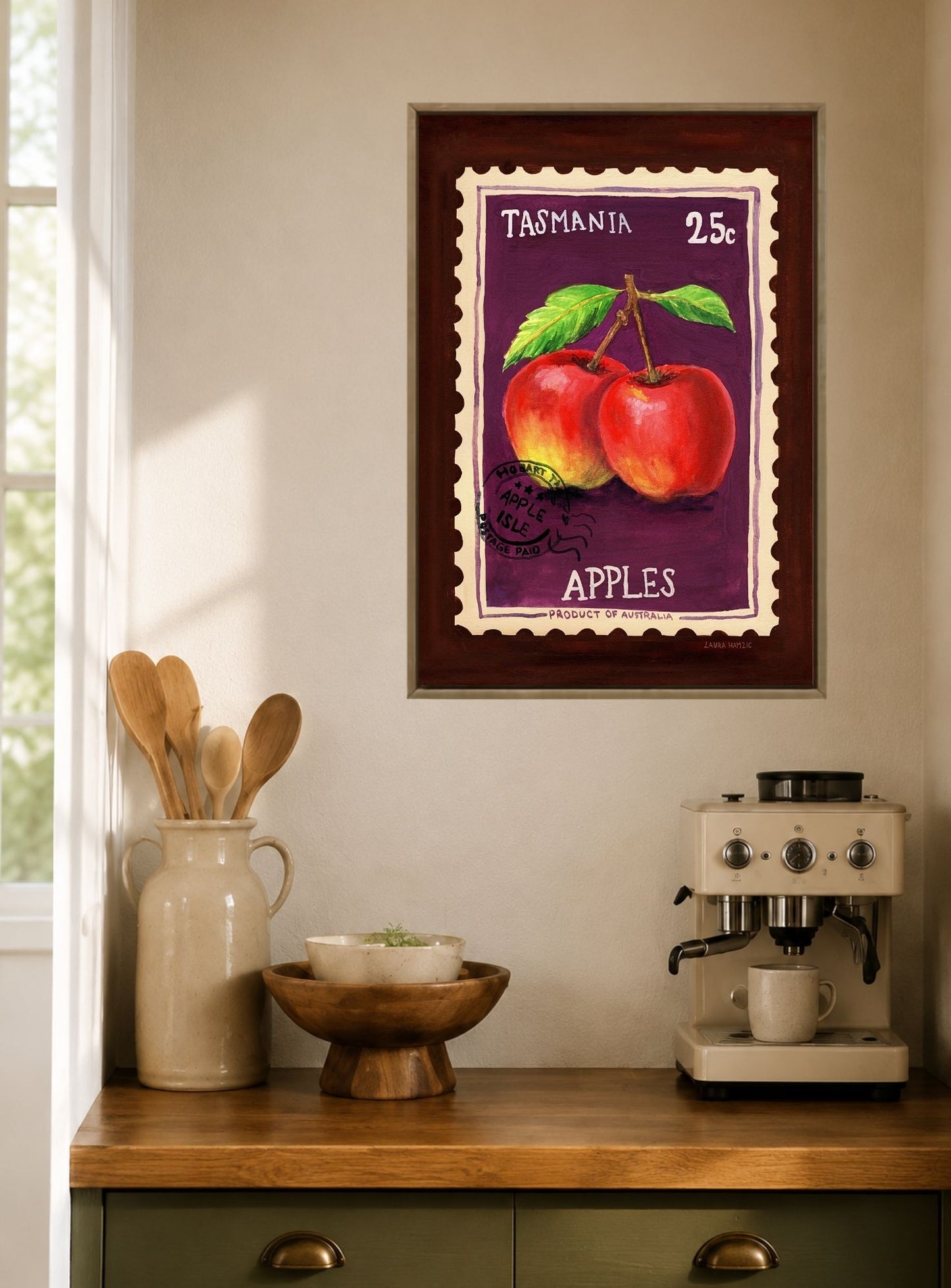 A framed vintage-style wall print featuring two red apples on a purple background with the words 'TASMANIA' and 'APPLES', displayed above a kitchen counter with a cream coffee machine, wooden bowl, and utensils.