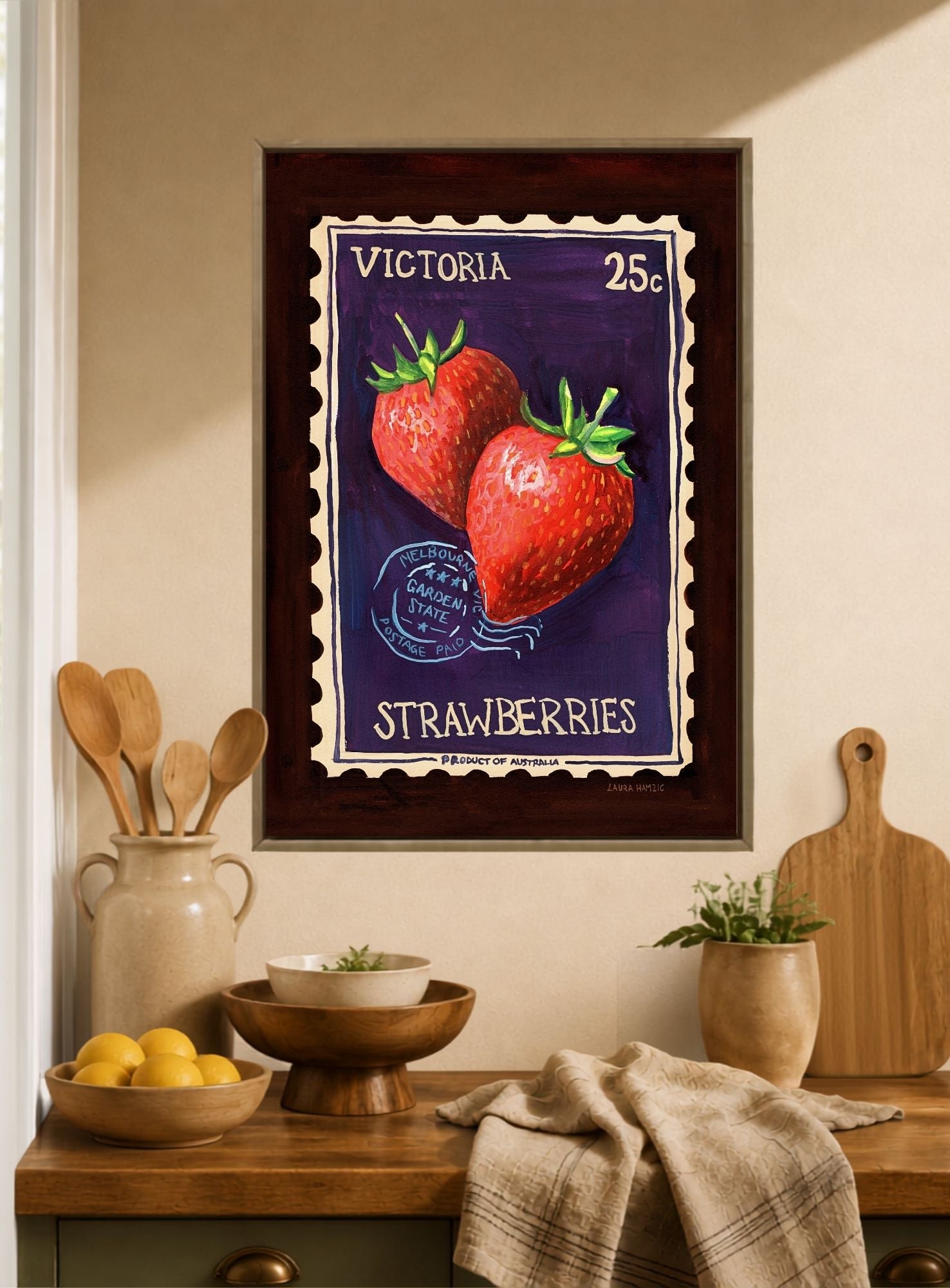 A framed vintage-style strawberry poster hangs on a beige wall above a wooden counter with kitchen items, including lemons, bowls, utensils, and a cutting board.