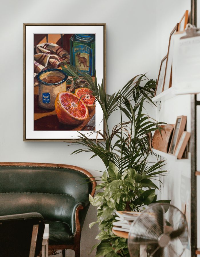 Framed still life painting of an everyday moment showing a kitchen bench with grapefruit, a freshly brewed cup of tea and a vintage bushells tea tin on a wall above a green chair.